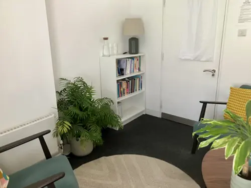 Small therapy room with white walls, dark grey carpet, 2 green armchairs, plants and a bookcase.